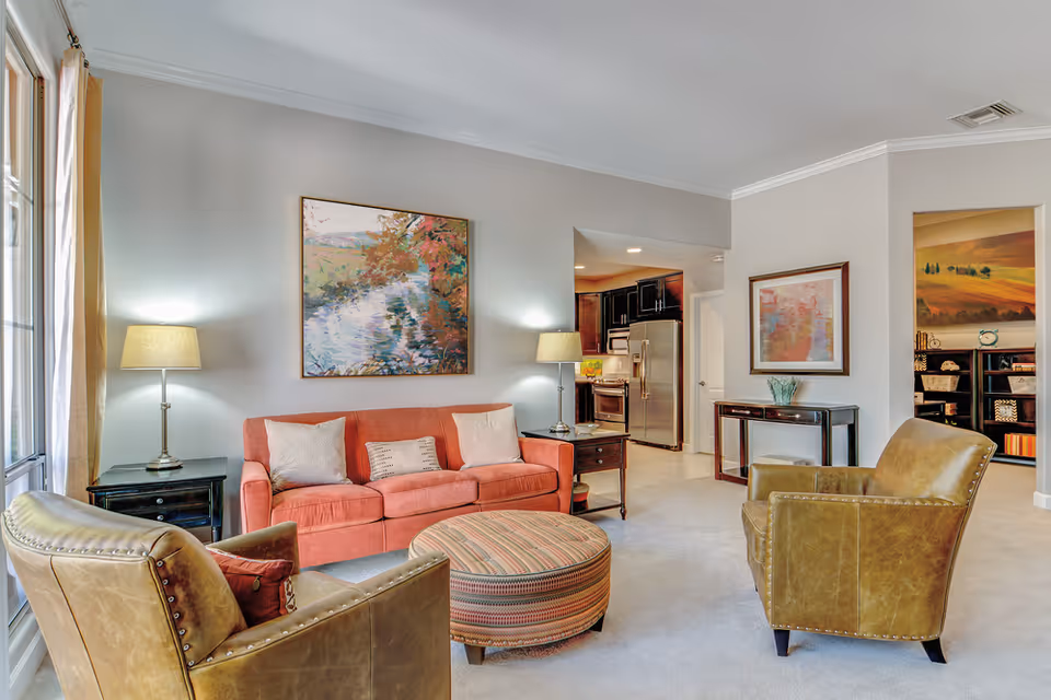A cozy living room featuring a coral-colored sofa with three cushions, two brown leather armchairs, and a round striped ottoman in the center. The room has light-colored walls adorned with two framed paintings, two side tables with lamps, and a large window with beige curtains letting in natural light. In the background, a kitchen area with stainless steel appliances and dark cabinetry is visible.
