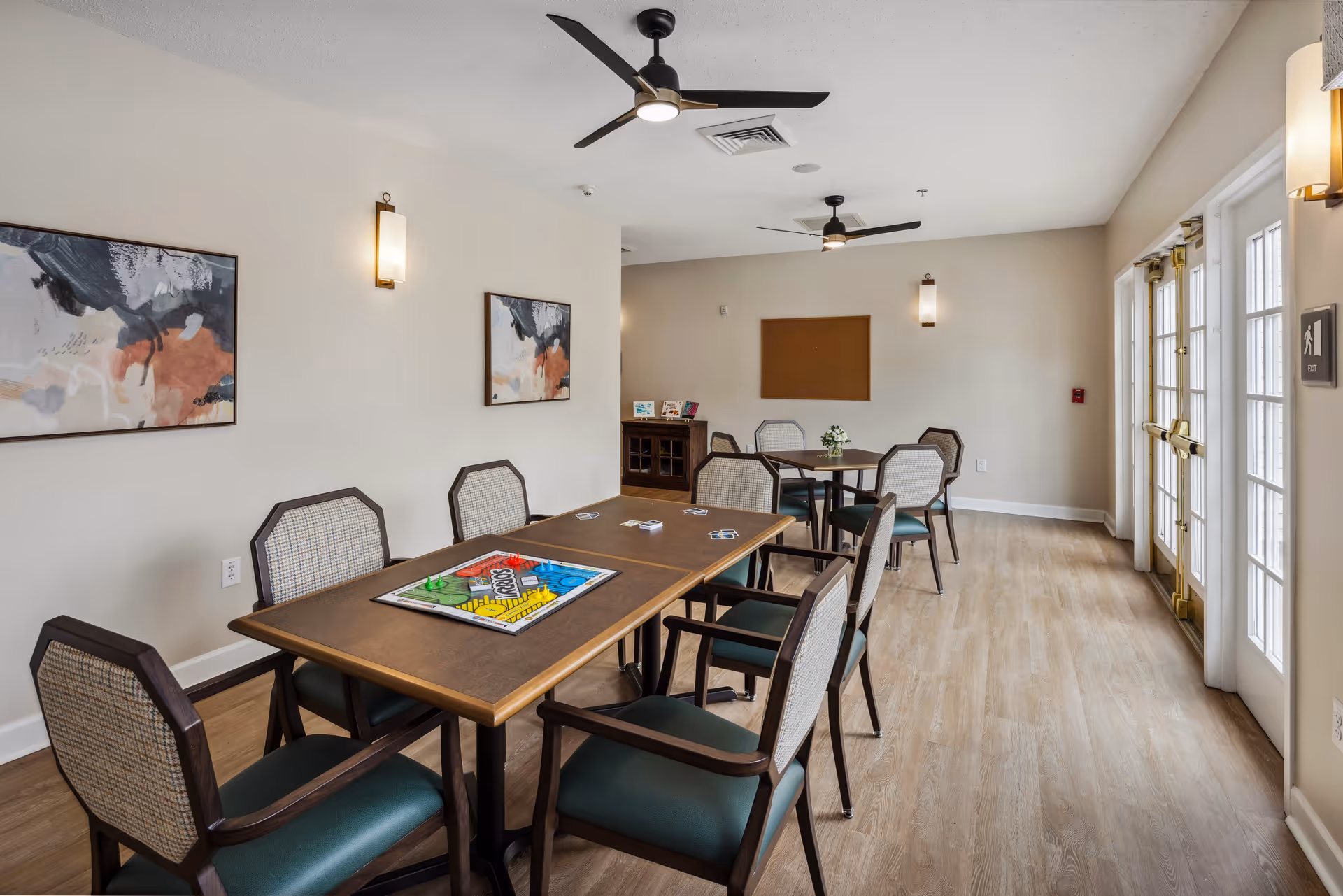 A well-lit common area with two tables and several chairs arranged around them. One table has a board game set up on it. The room has light-colored walls with two abstract paintings, ceiling fans, wall sconces, and large glass doors letting in natural light.