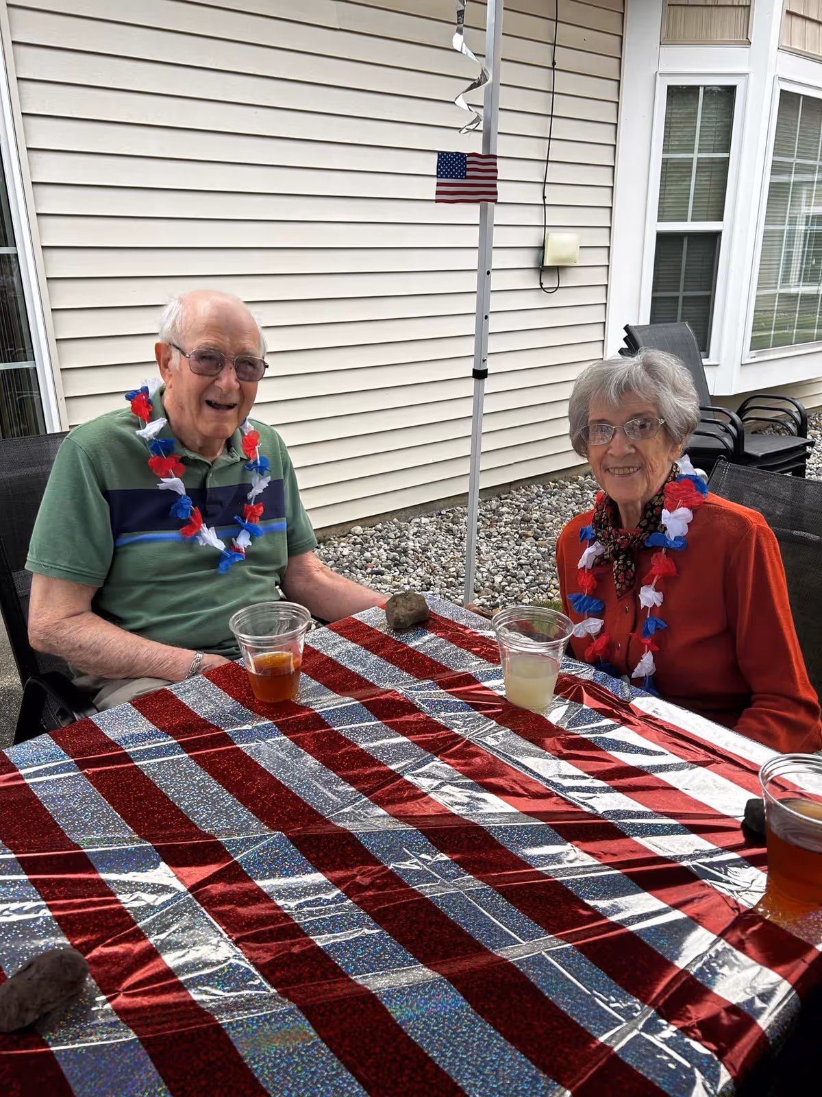Two elderly people sitting at a table outdoors with a red, white, and blue striped tablecloth. Both are wearing red, white, and blue leis. There is a small American flag attached to a pole behind them. The setting appears to be a patio area next to a building with beige siding and windows.