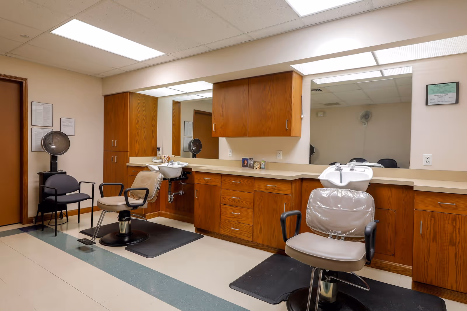 Interior view of a senior living facility's hair salon area featuring two salon chairs with protective covers, two hair washing sinks, wooden cabinets, large mirrors, and a couple of black chairs against the wall. The room has a tiled floor and fluorescent ceiling lights.