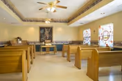 Interior view of a small chapel or meditation room with wooden pews arranged in rows facing a small altar at the front. The room has light-colored walls, two stained glass windows on the right side, a ceiling fan with lights, and a framed religious artwork above the altar.