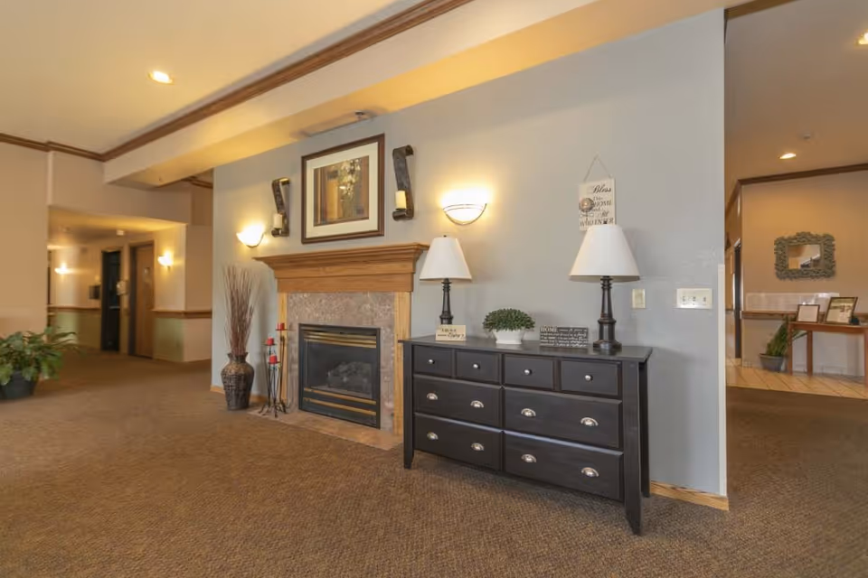 Carpeted interior lobby with a fireplace and mantel beside a black dresser topped with lamps and decorative items.