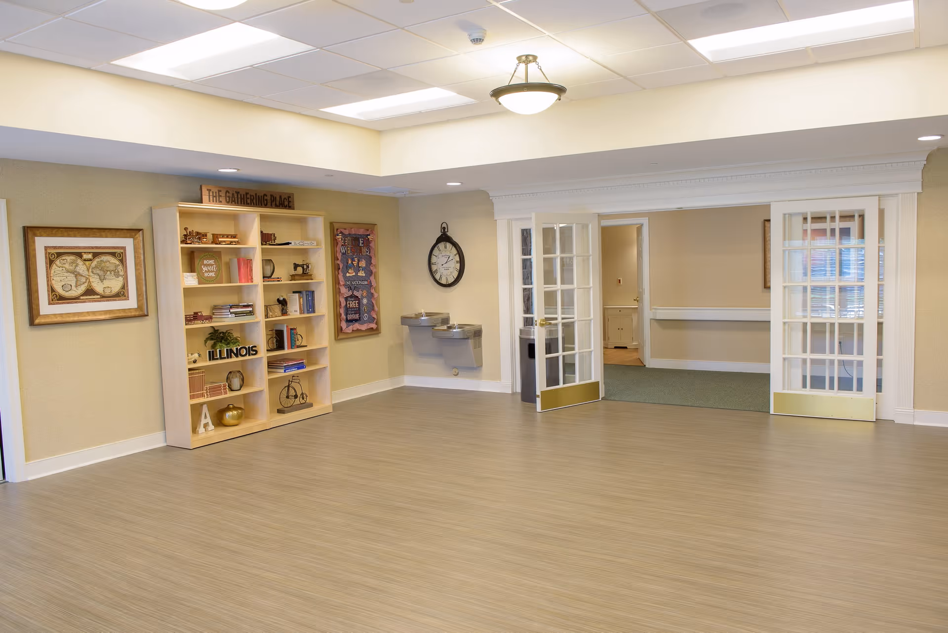 A spacious, well-lit common area with light wood flooring and beige walls. On the left side, there is a wooden bookshelf filled with decorative items and books, with a sign on top that reads 'The Gathering Place.' Above the bookshelf is a framed world map. A wall clock and two water fountains are mounted on the far wall. To the right, there are double glass doors with white frames leading to another room with carpeted flooring.