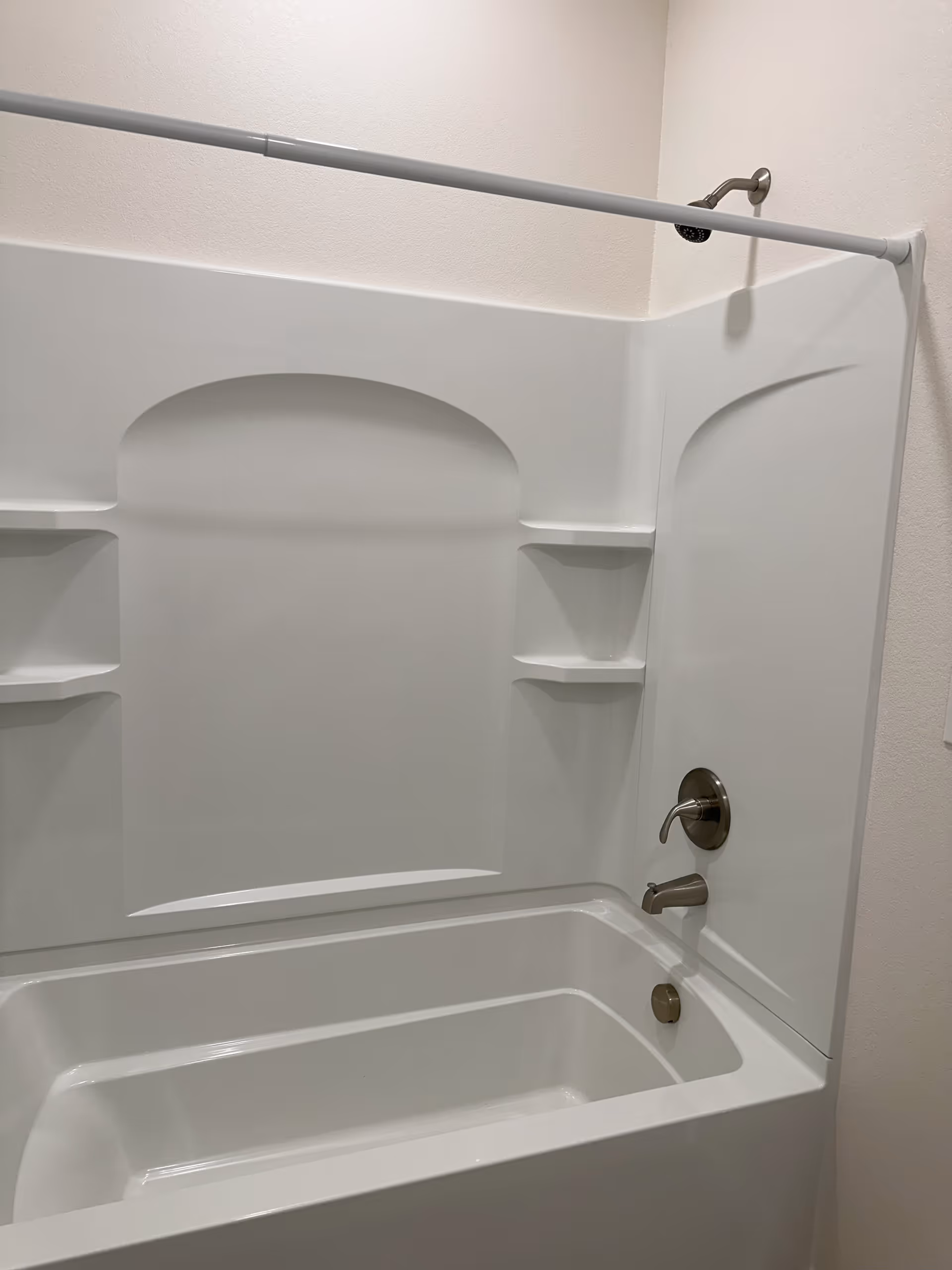 A clean white bathtub with built-in shelves and a silver faucet and showerhead mounted on the wall. A white shower curtain rod is installed above the bathtub.