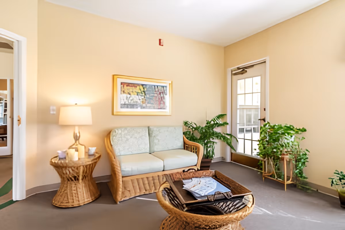A cozy sitting area with a wicker loveseat with light green cushions, a matching wicker side table with a lamp and candles, and a wicker coffee table with a tray and papers. There are several green plants near a glass door letting in natural light, and a framed abstract painting on the beige wall.
