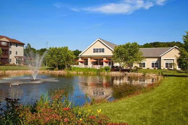A scenic view of Monroe Village featuring a pond with a water fountain, surrounded by green grass and colorful flowers. In the background, there are two buildings, one with a beige exterior and large windows, and the other a multi-story building with balconies. The sky is clear and blue.