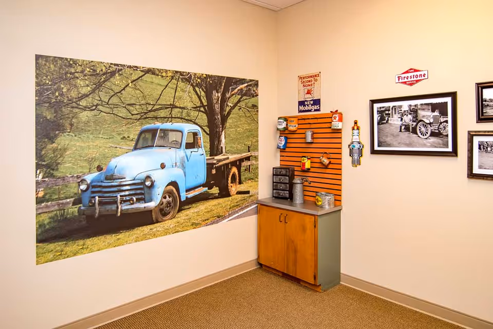 Corner of an interior room with a large mural of a blue vintage truck, a small cabinet topped with vintage gas-themed decor and framed black-and-white photos on the wall.