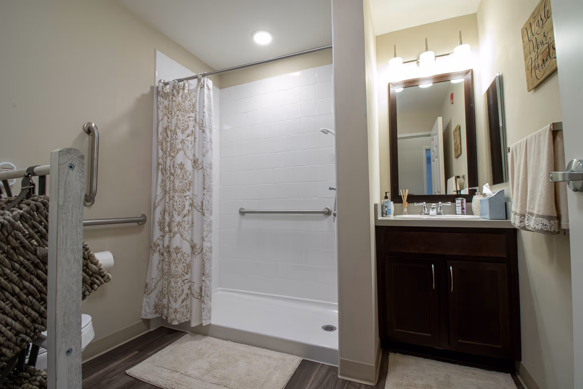 A clean and well-lit bathroom featuring a walk-in shower with a white tiled wall and a decorative shower curtain. There is a dark wood vanity with a sink, a large mirror above it, and a light fixture with three lights. The bathroom also has grab bars for accessibility, a towel rack with a beige towel, and a beige bath mat on the floor.