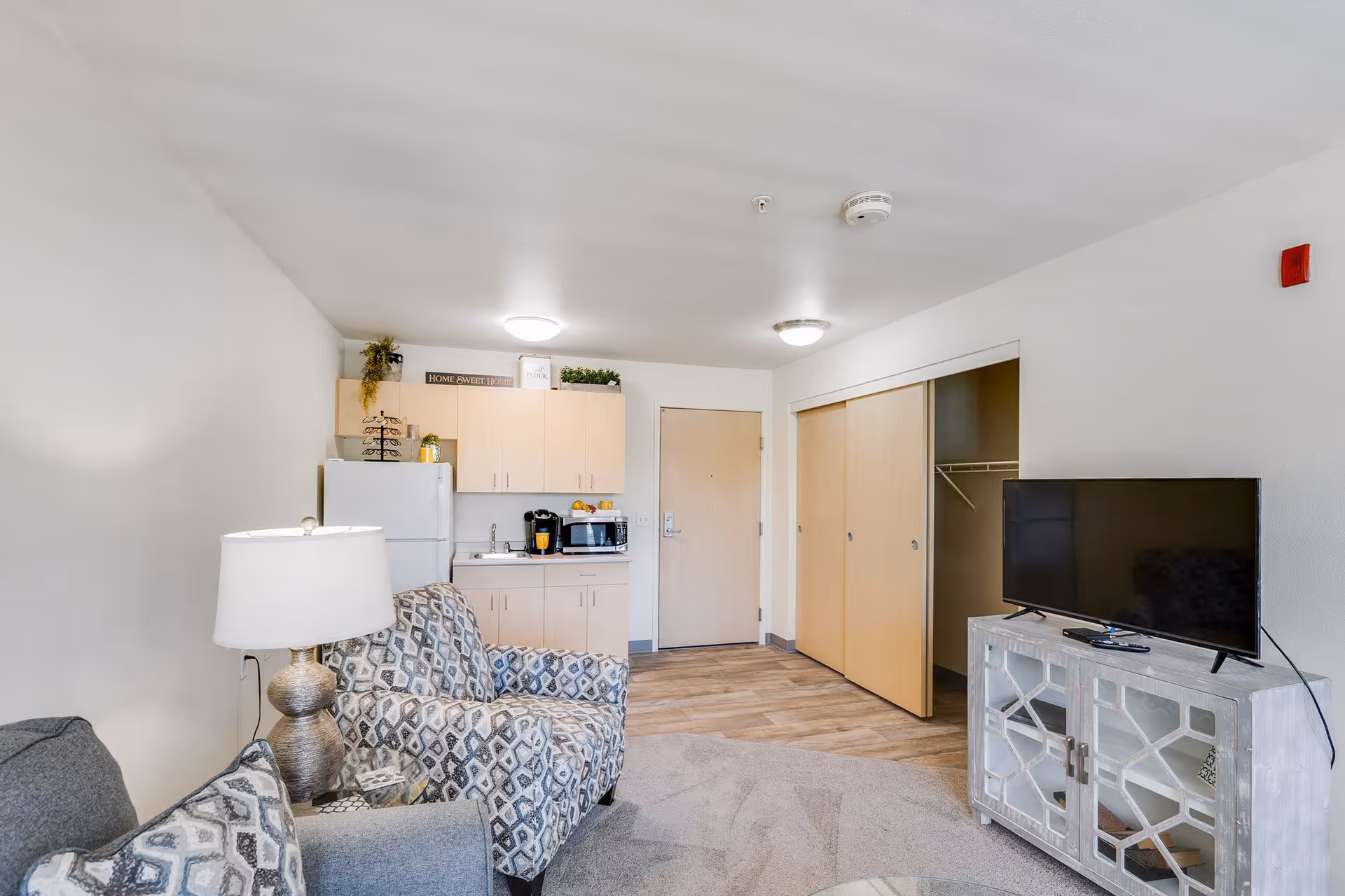 Interior view of a small living space in an independent living facility. The room features a patterned armchair, a lamp, a small kitchenette with a refrigerator, microwave, and cabinets, a TV on a decorative stand, and a closet with sliding doors. The flooring is a combination of carpet and wood, and the walls are painted white.