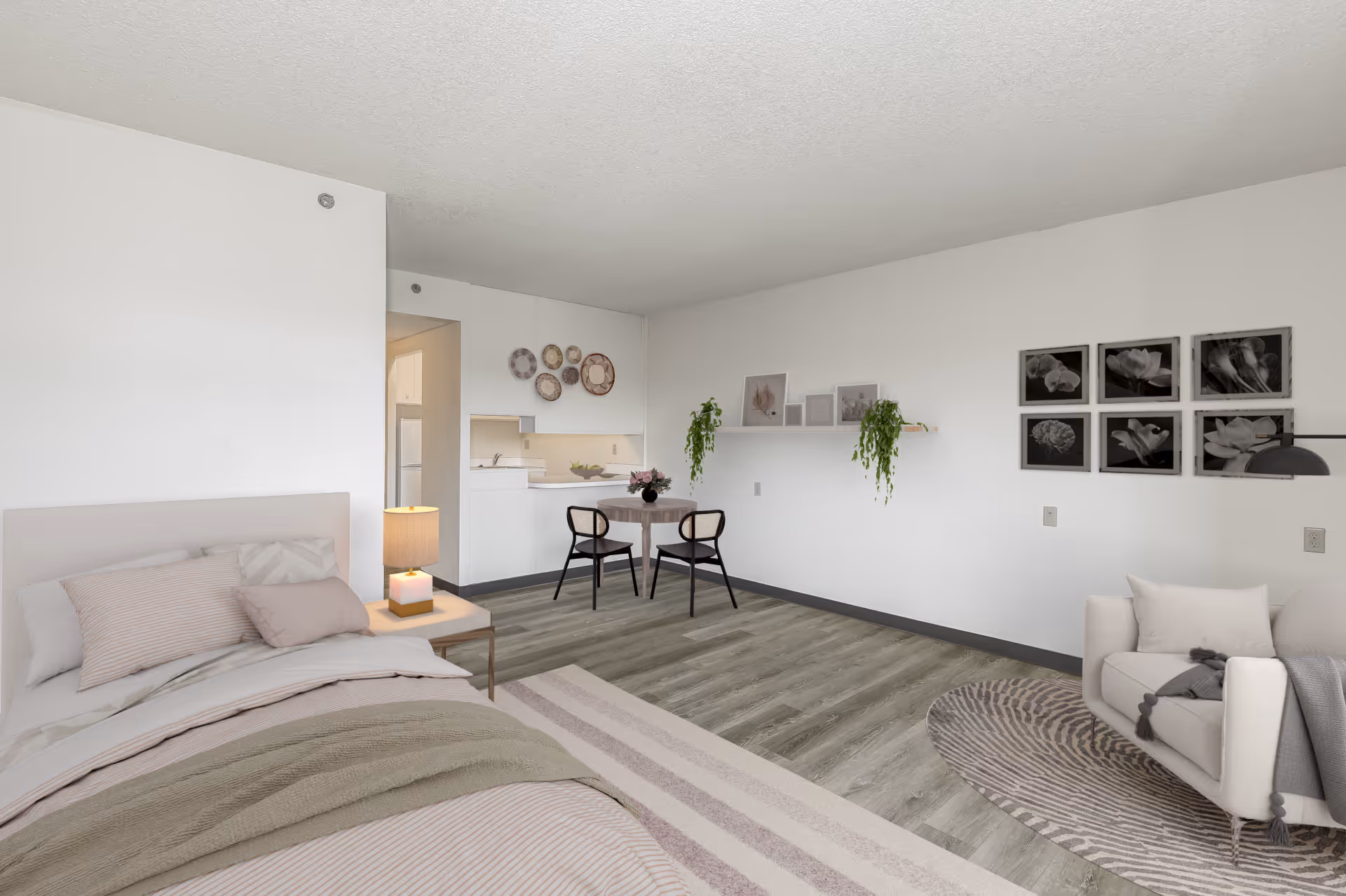A bright, modern studio apartment featuring a bed with neutral-colored bedding on the left, a small dining table with two chairs in the center near a kitchenette, and a cozy armchair with a throw blanket on the right. The walls are decorated with framed black and white floral artwork and hanging plants. The flooring is wood-style laminate with a striped area rug near the bed and a round patterned rug near the armchair.