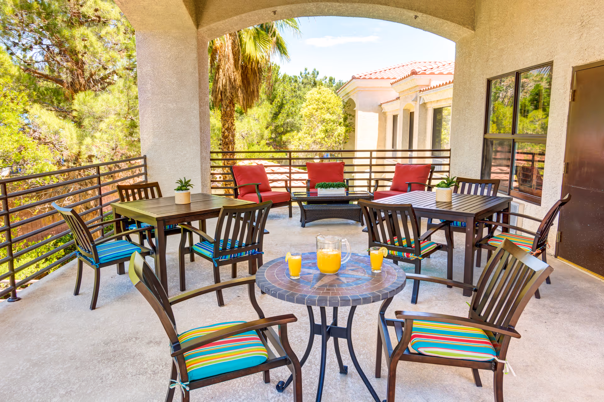 Outdoor covered patio area with multiple wooden tables and chairs featuring colorful striped cushions. There are two red cushioned armchairs and a coffee table with a small plant. A small round table in the foreground holds a pitcher of orange juice and two glasses. The patio overlooks green trees and a building with a tiled roof.