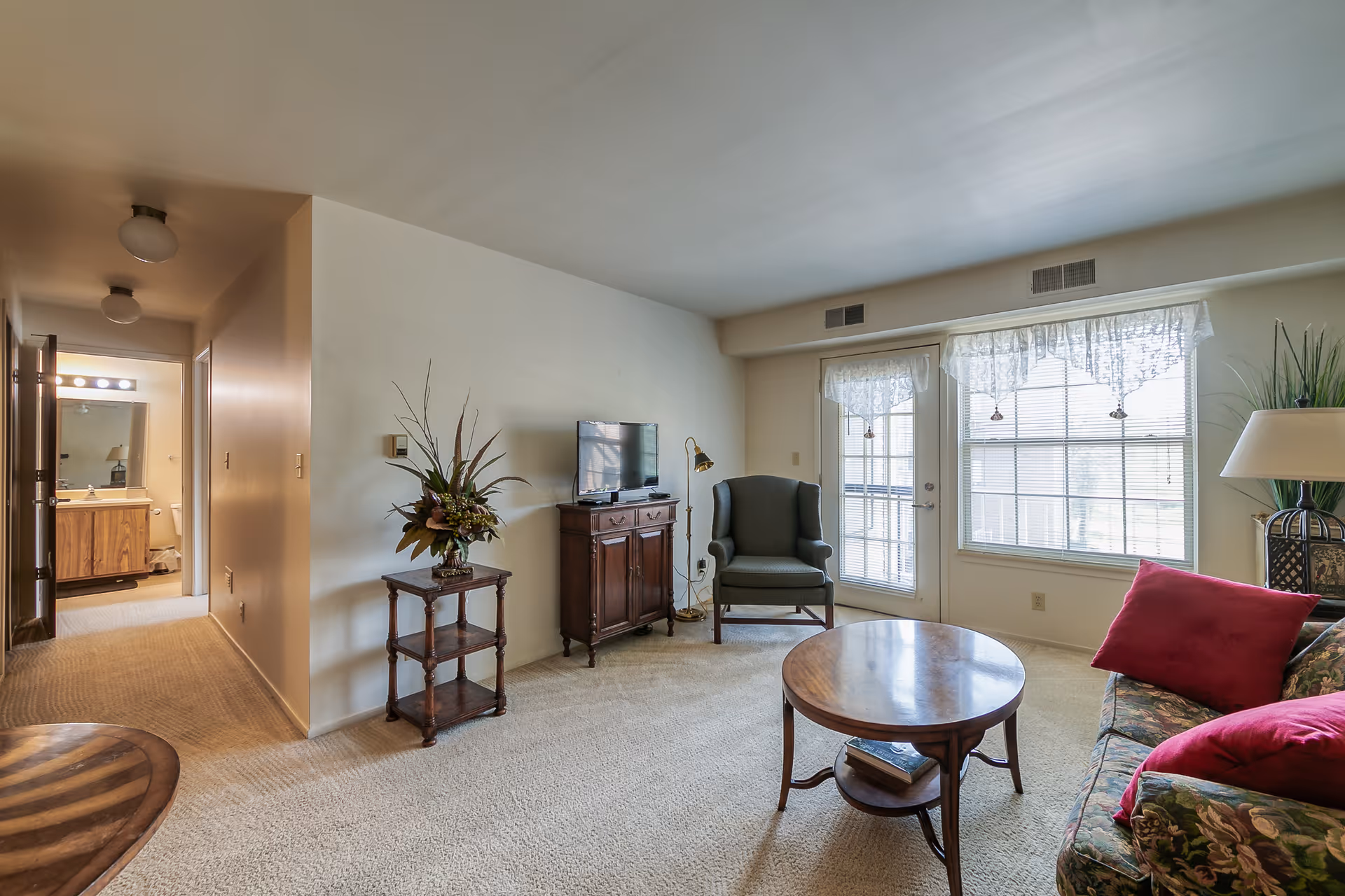 A cozy living room with beige carpet and walls, featuring a floral-patterned sofa with red cushions, a round wooden coffee table with a book underneath, a green armchair next to a floor lamp, a wooden TV stand with a flat-screen TV, and a small wooden shelf with a decorative plant. Large windows with lace curtains and a glass door allow natural light to fill the room. A hallway leading to a bathroom is visible on the left side.