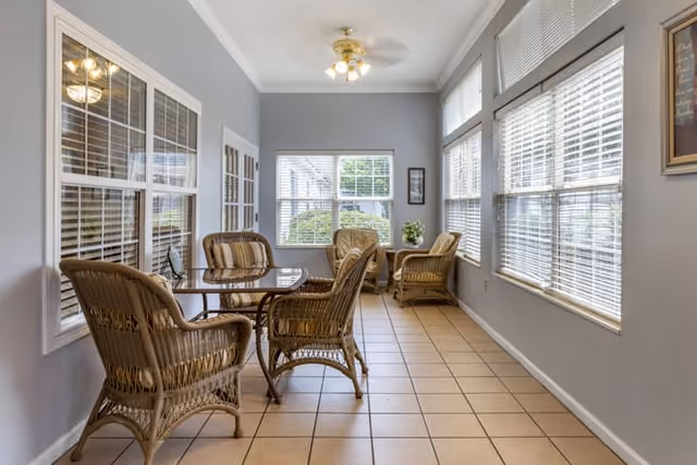 A bright sunroom with large windows on two sides, featuring a ceiling fan with lights, a glass-top table surrounded by four wicker chairs, and two additional wicker armchairs near the window. The floor is tiled, and the walls are painted light gray.
