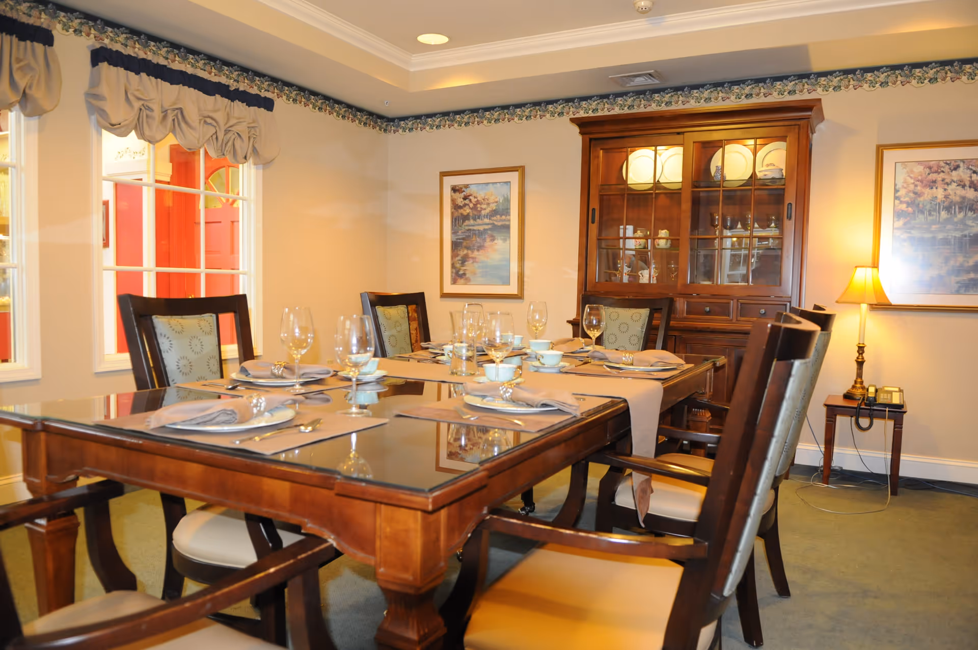 A formal dining room with a wooden table set for six people, featuring placemats, napkins, plates, cups, and wine glasses. The room has upholstered chairs, a wooden china cabinet displaying dishes, framed artwork on the walls, a table lamp on a side table, and windows with decorative valances.