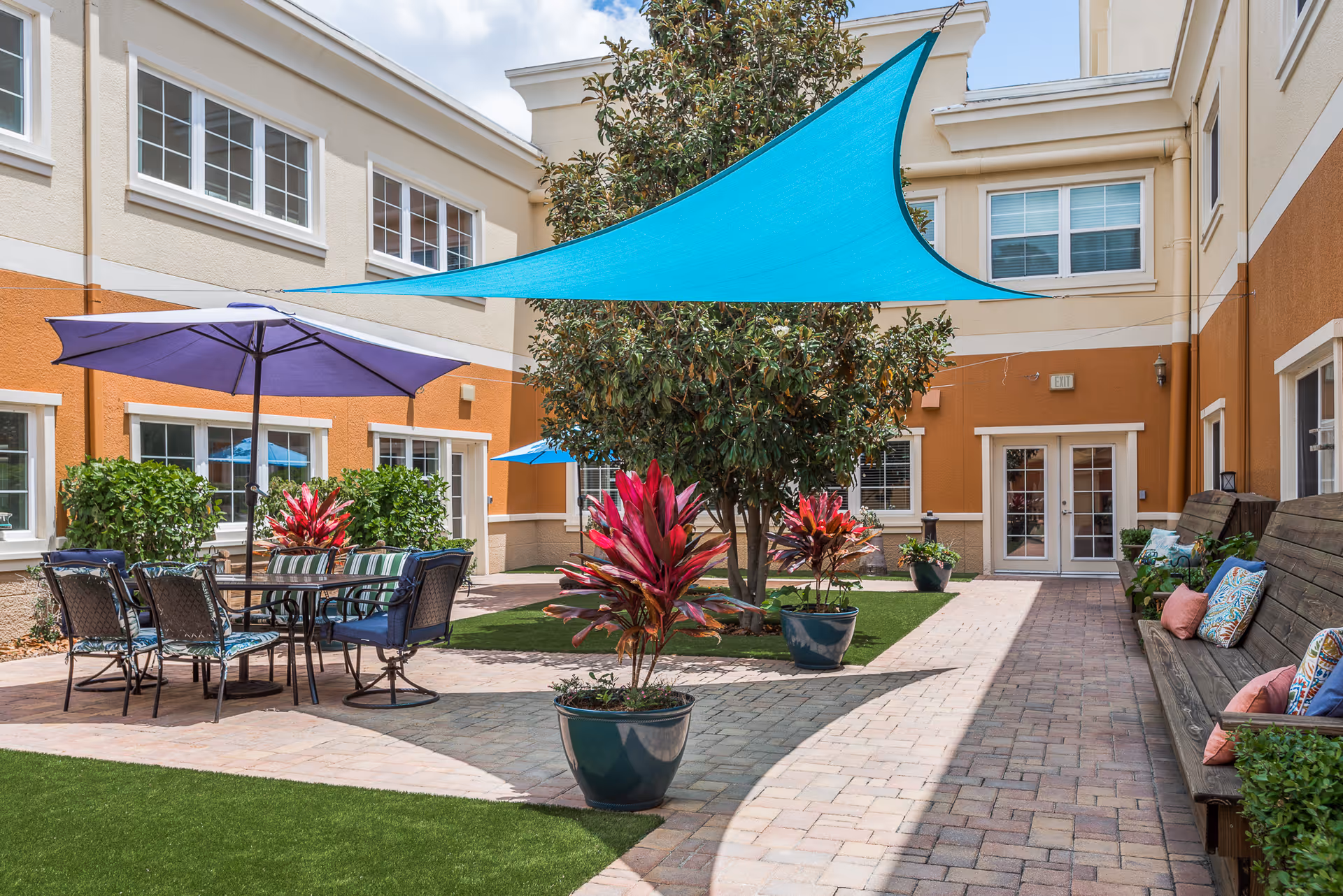 Outdoor courtyard area with a paved walkway, green artificial grass, potted plants with red leaves, a large tree, a blue triangular shade sail overhead, a purple patio umbrella shading a table with six chairs, and wooden benches with colorful cushions along the right side. The courtyard is surrounded by a beige and orange building with multiple windows and a double door exit.