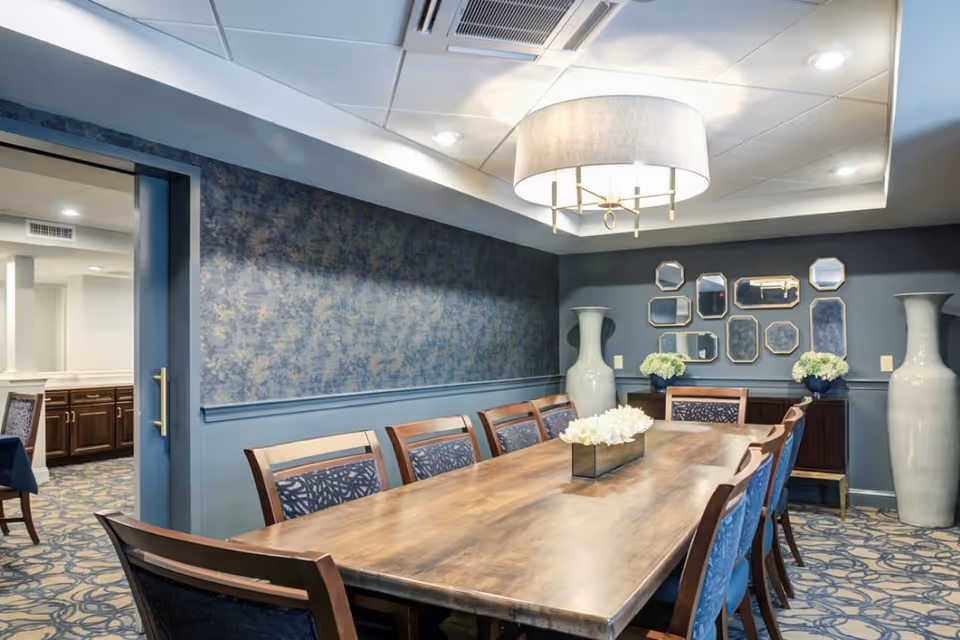A well-lit dining room with a long wooden table surrounded by wooden chairs with patterned cushions. The walls are painted blue with a decorative wallpaper accent and a collection of small mirrors arranged on one wall. Two large white vases with floral arrangements are placed on either side of a wooden sideboard. A modern ceiling light fixture hangs above the table. The room has a patterned carpet and an open doorway leading to another room with cabinetry.