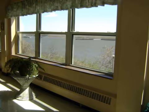 Sunlit interior with wide windows overlooking water, and a potted plant by the radiator.