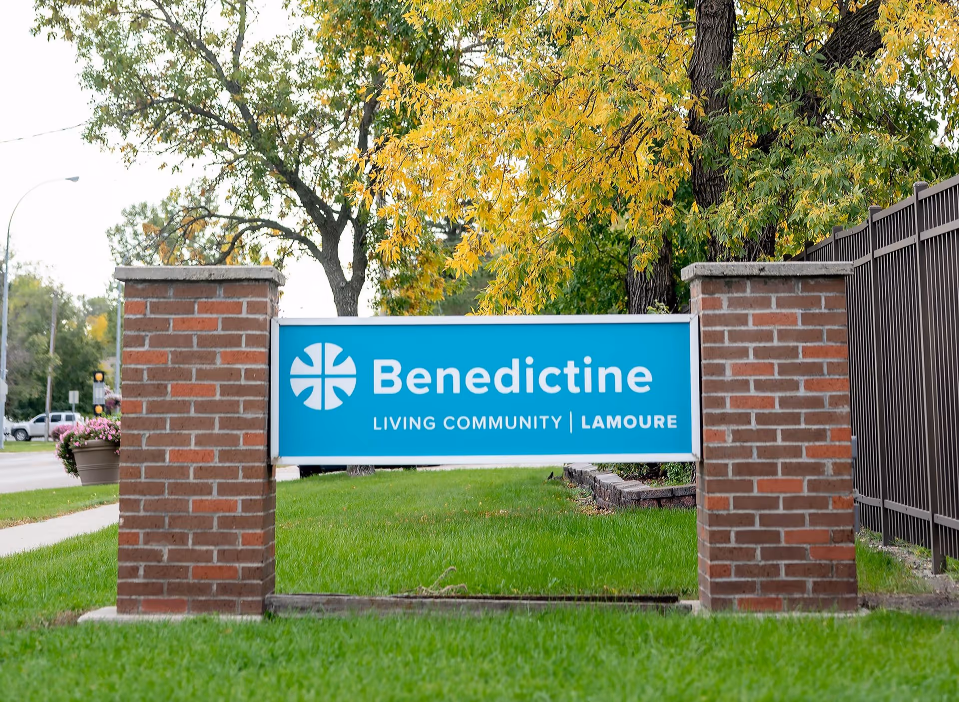 A blue sign with white text reading 'Benedictine Living Community | LaMoure' mounted between two brick pillars on a grassy area with trees and a sidewalk nearby.