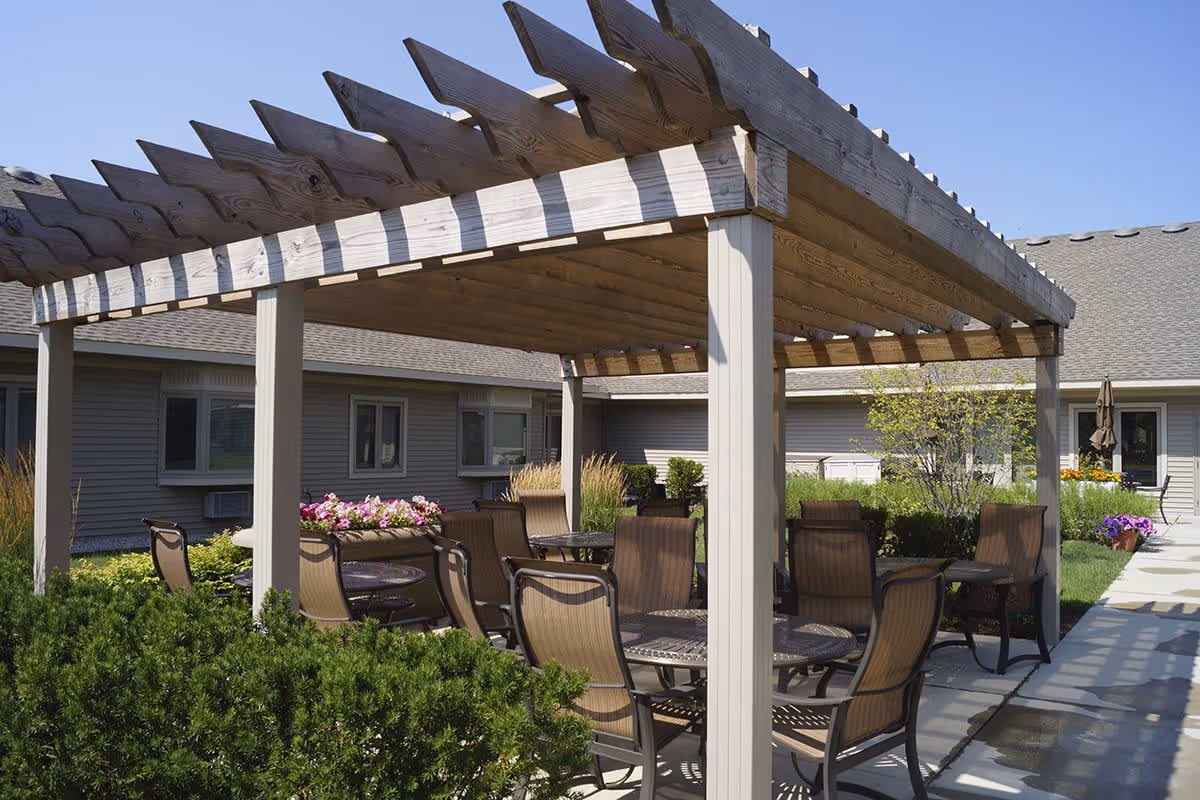 Outdoor seating area at Linden Square Senior Care featuring a wooden pergola with multiple tables and chairs underneath, surrounded by greenery and flowers, with the building visible in the background under a clear blue sky.