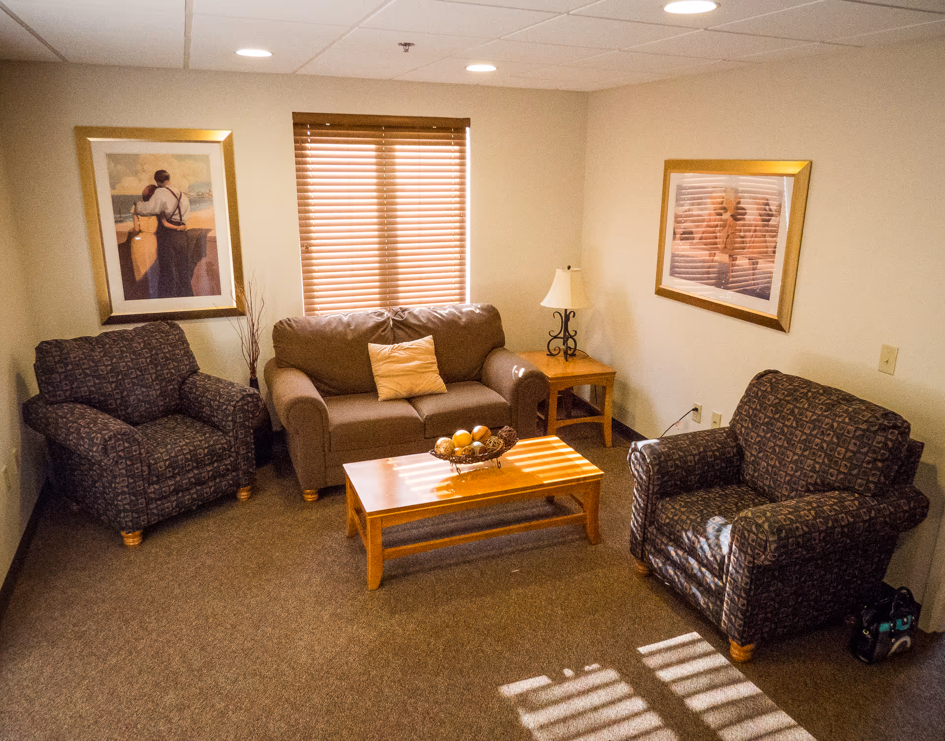 A cozy living room with a brown loveseat, two patterned armchairs, a wooden coffee table with a decorative bowl, a side table with a lamp, two framed paintings on the walls, and a window with wooden blinds letting in sunlight.