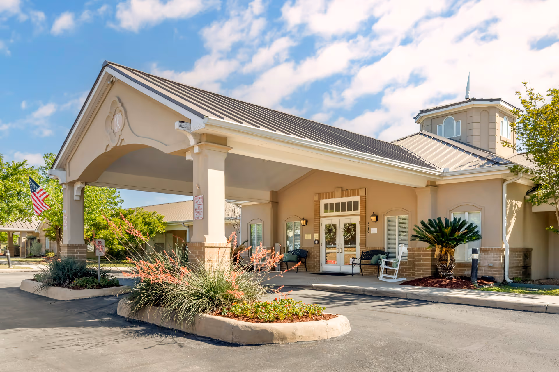 Entrance of a senior living facility with a covered drop-off area, beige exterior walls, and a metal roof. There are benches and a rocking chair near the glass double doors, landscaped plants in front, and an American flag on the left side. The sky is partly cloudy.