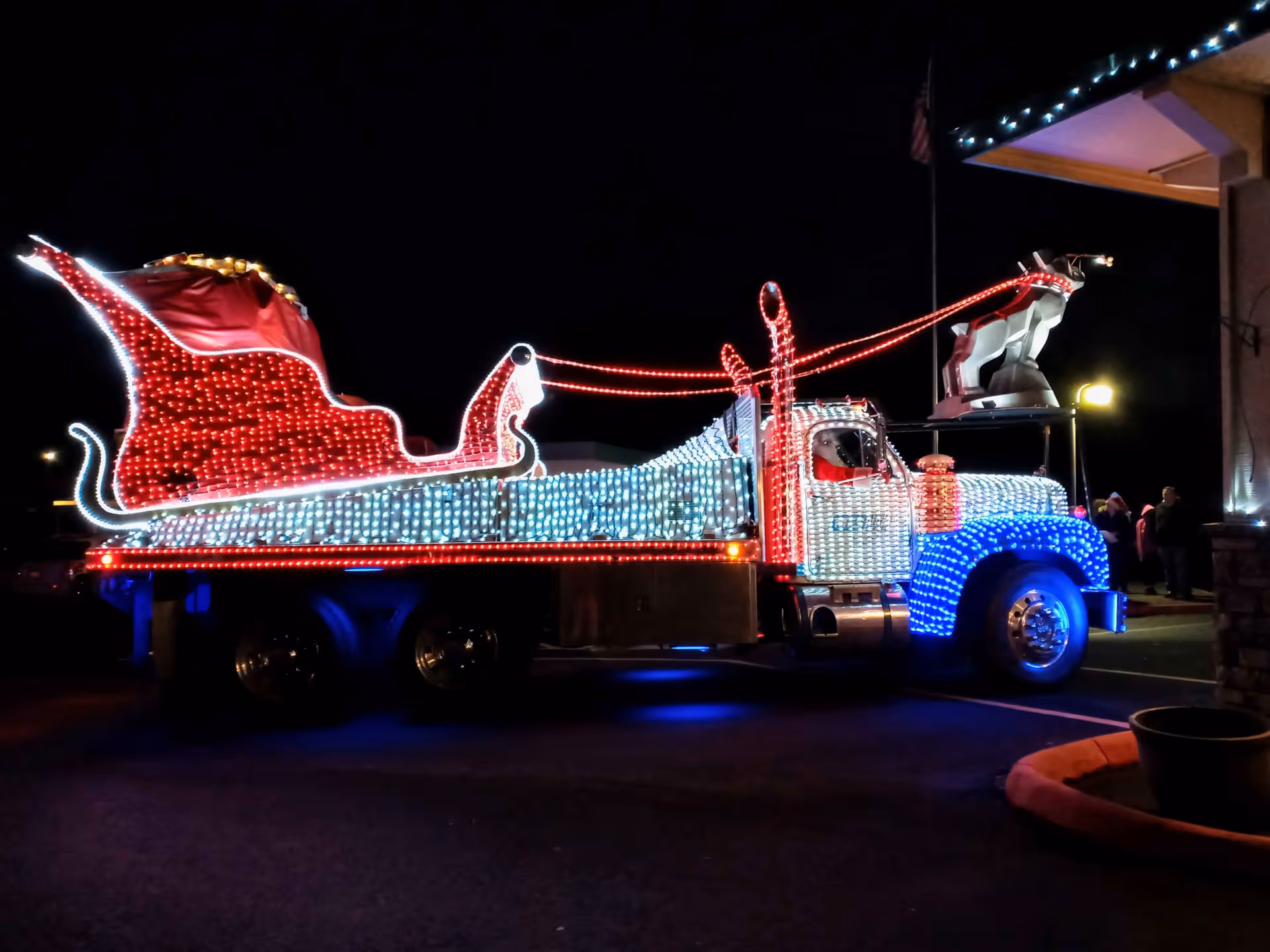 A large truck decorated with colorful Christmas lights at night, featuring a red and white sleigh on the back and a reindeer figure mounted on the front. The truck is parked near a building with a few people visible in the background.
