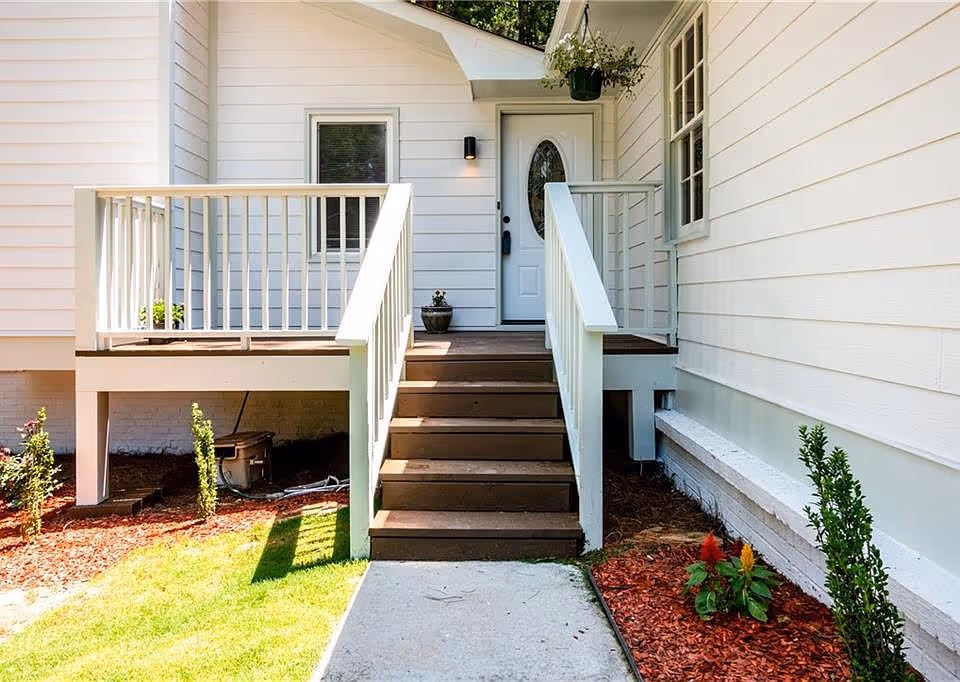 Front entrance of a white house with a small wooden porch, stairs leading up to a white door with an oval glass window, surrounded by green plants and mulch landscaping.