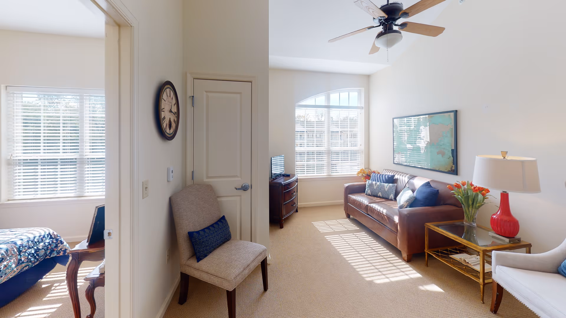 A bright and cozy living room with a brown leather sofa adorned with decorative pillows, a glass-top side table with a red lamp and a vase of flowers, a white armchair, and a ceiling fan. Adjacent to the living room is a bedroom with a bed covered in a patterned quilt, visible through an open doorway. The room has large windows with white blinds allowing natural light to fill the space.