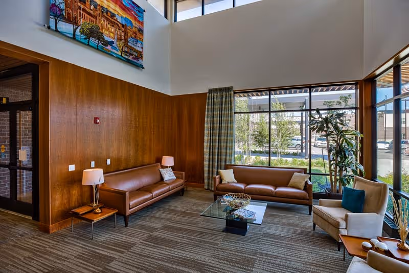 A bright and spacious senior living facility common area with large windows letting in natural light. The room features two brown leather sofas, a beige armchair with a blue cushion, a glass coffee table with decorative items, two side tables with lamps, and a large colorful painting on the upper wall. Outside the windows, trees and parked cars are visible.