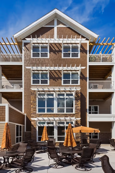 Outdoor patio area with multiple round tables and chairs, each table shaded by an orange umbrella, in front of a multi-story building with brown siding, white trim, and balconies under a blue sky.