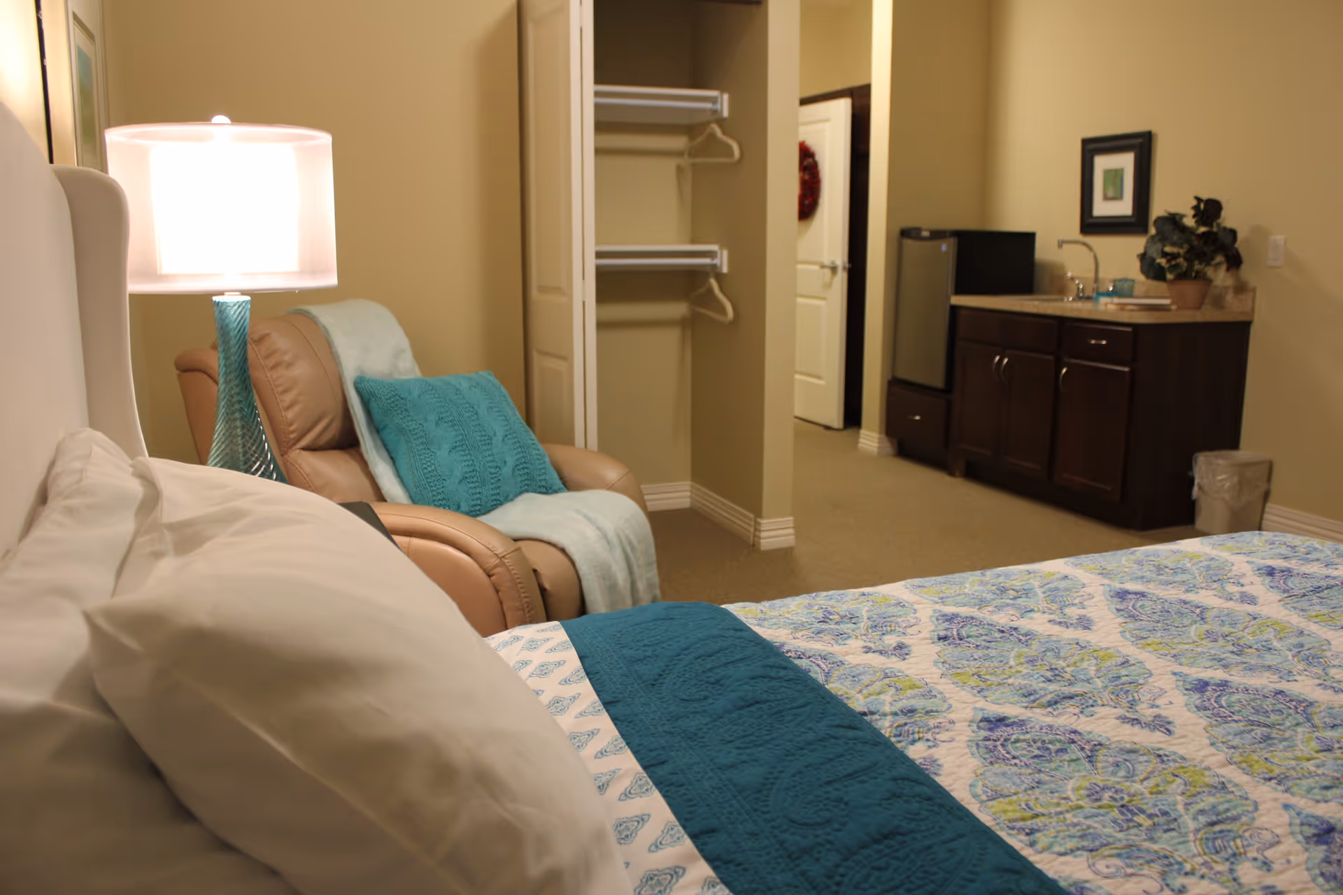 A cozy bedroom in a senior living facility featuring a bed with white pillows and a patterned blue and green quilt. Next to the bed is a beige recliner chair with a teal pillow and matching throw blanket. A blue glass table lamp is on a nightstand beside the chair. In the background, there is an open closet with hangers, a small kitchenette area with a sink, countertop, mini fridge, and a potted plant. The walls are painted beige and the floor is carpeted.
