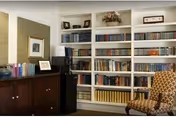 Interior view of a room with a large white bookshelf filled with books and decorative items. To the left, there is a dark wooden cabinet with framed pictures and a lamp on top. A patterned upholstered chair is partially visible on the right side of the image.