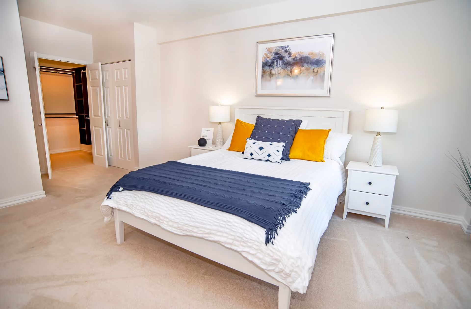 A bright bedroom with a white bed frame and bedding, accented by a navy blue throw and decorative pillows in navy and yellow. Two white nightstands with matching lamps flank the bed. A framed artwork hangs above the bed, and an open closet with white doors is visible to the left.