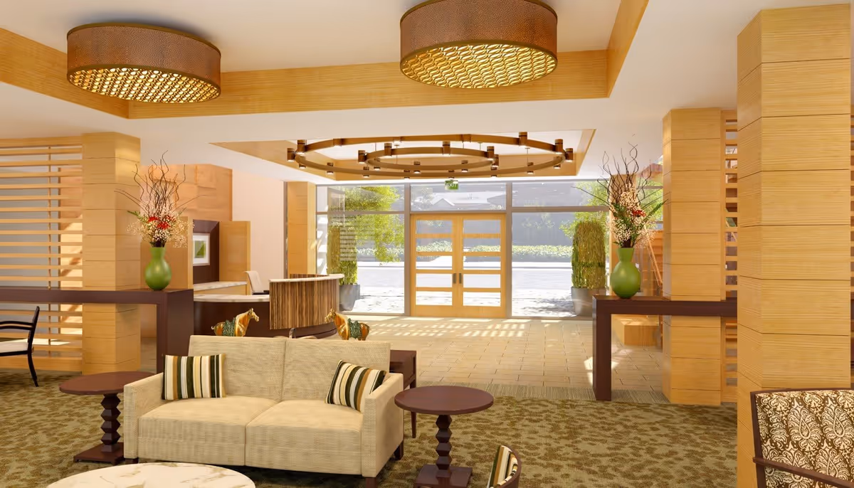 A bright and spacious lobby area with beige sofas, wooden tables, and decorative vases with flowers. Large glass doors at the back let in natural light, and the ceiling features modern circular light fixtures. The space has a warm, inviting atmosphere with wood paneling and patterned carpet.