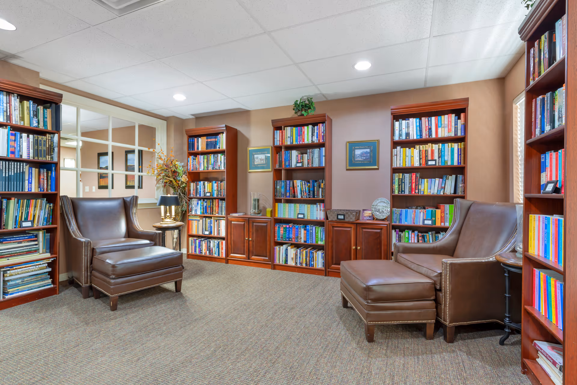 A cozy reading room with multiple wooden bookshelves filled with books, two brown leather armchairs, one with an ottoman, a small side table with a lamp, and framed pictures on the wall.
