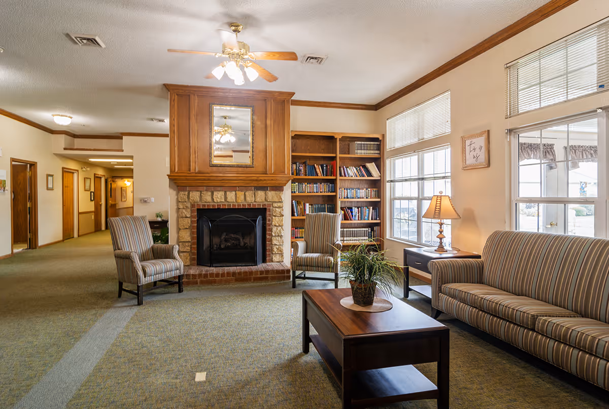 Bright communal living room with a stone fireplace, bookshelves, striped sofa and chairs, coffee table, and large windows.