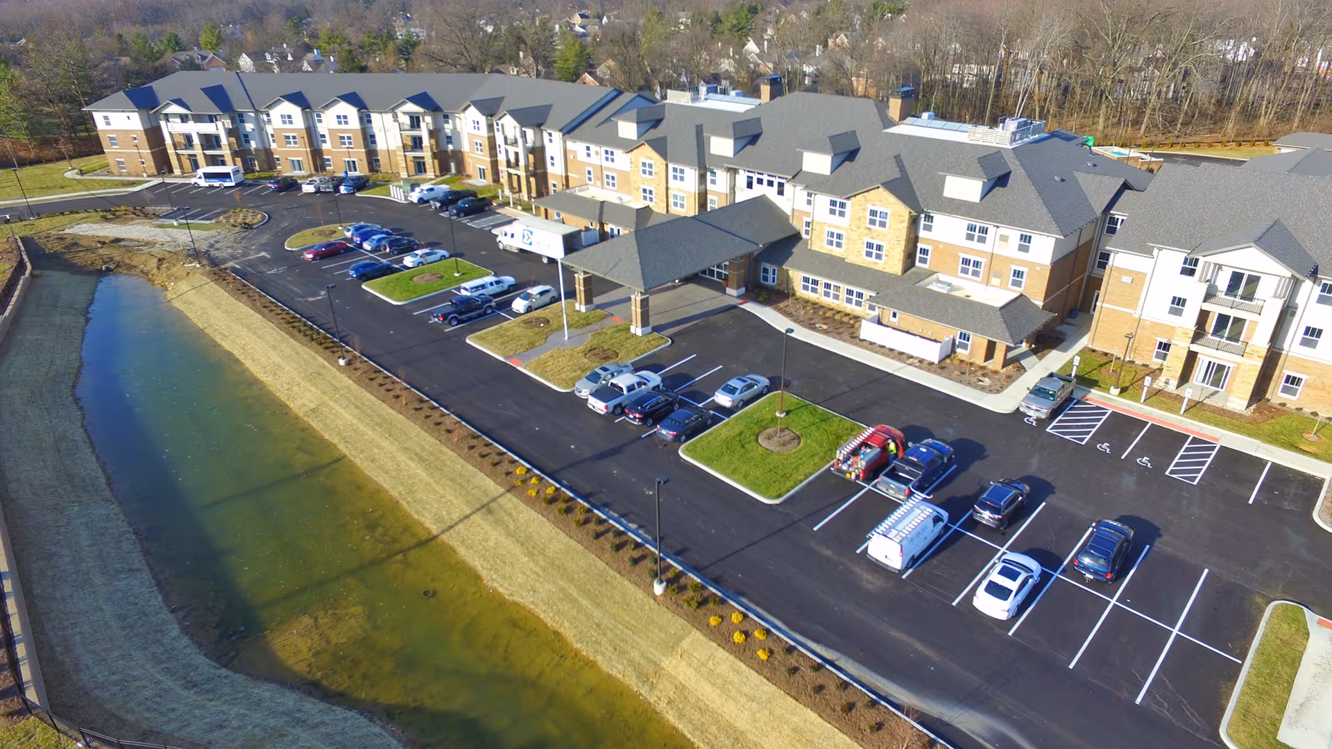 Aerial view of Deerfield Springs Retirement Resort showing a large three-story building with a covered entrance, multiple parked cars in the parking lot, landscaped grassy areas, and a narrow water body running alongside the property.