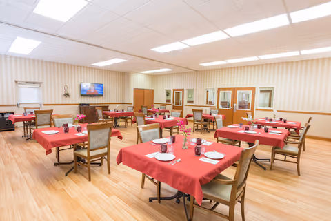 A spacious dining room with multiple tables covered in red tablecloths, each set with plates, cups, and utensils. The room has light wood flooring, striped wallpaper, and fluorescent ceiling lights. There is a television mounted on the far wall and several chairs arranged around the tables.