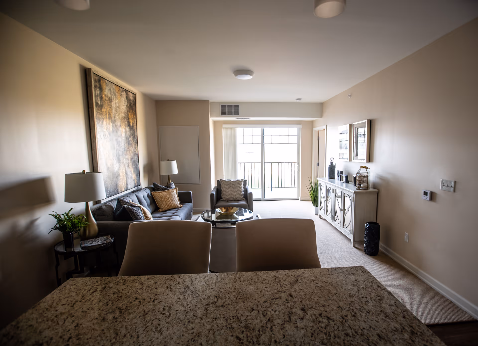 A cozy living room with a gray sofa adorned with cushions, a matching armchair, a glass coffee table, and a side table with a lamp and plant. There is a large abstract painting on the wall behind the sofa. The room has beige walls and carpet, a white sideboard with decorative items, and a sliding glass door leading to a balcony.