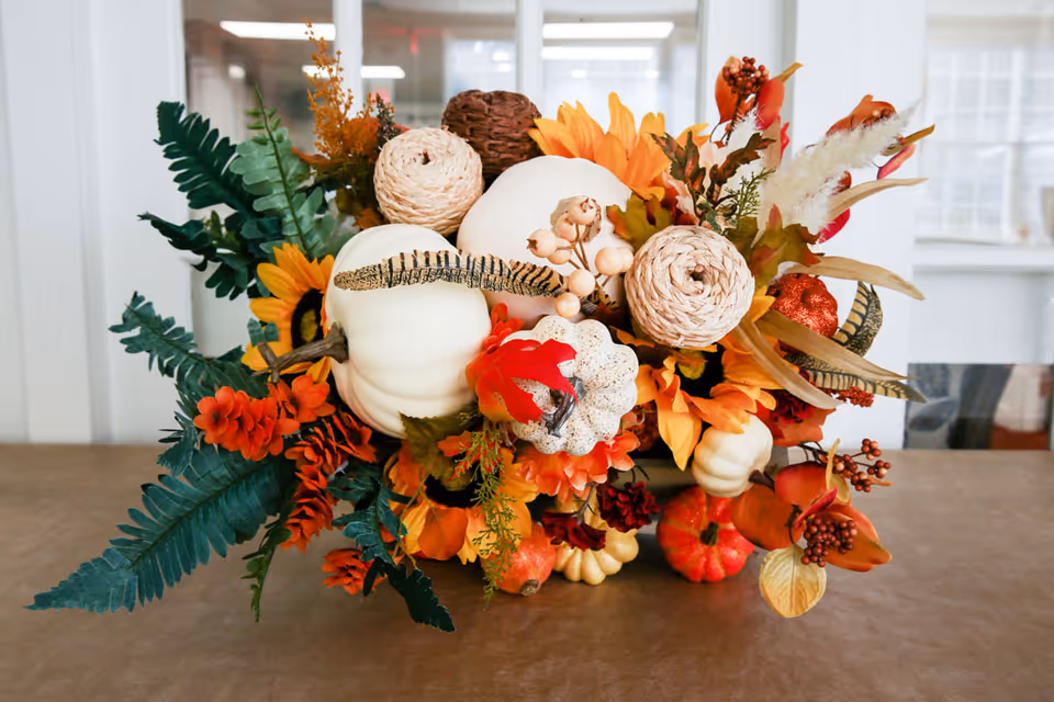 A decorative autumn-themed centerpiece featuring white pumpkins, orange flowers, green leaves, and various fall foliage arranged on a table with a blurred interior background.