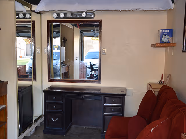 A small interior room with a dark wooden desk featuring multiple drawers, a large mirror above the desk, and a red cushioned chair or sofa to the right. There is a mirrored closet door on the left side reflecting part of the room and a window showing a parked car outside. A small wooden shelf on the wall holds a bingo game box.