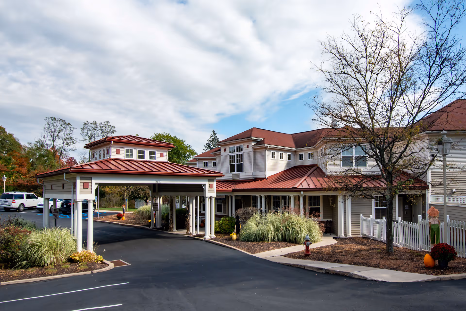Exterior view of Richland Woods by New Perspective senior living facility showing a covered entrance with white columns and a red roof. The building has beige siding and multiple windows. There is a parking lot with several cars, landscaped areas with bushes, a tree with no leaves, and autumn decorations including pumpkins and scarecrows.