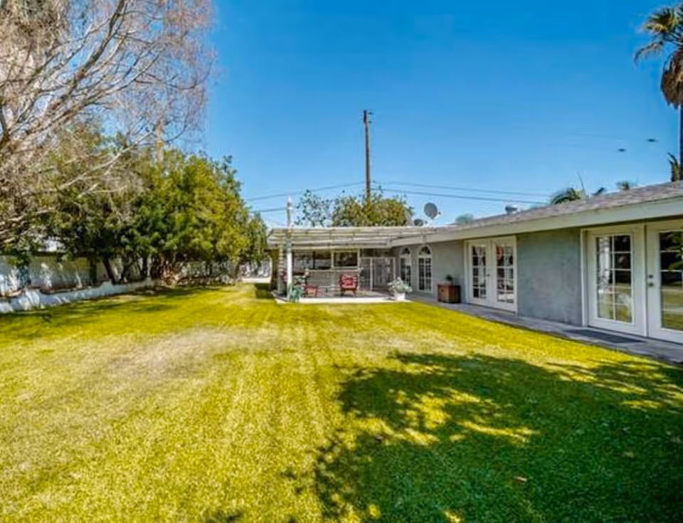 Wide grassy backyard with a covered patio attached to a single-story building under a clear blue sky.