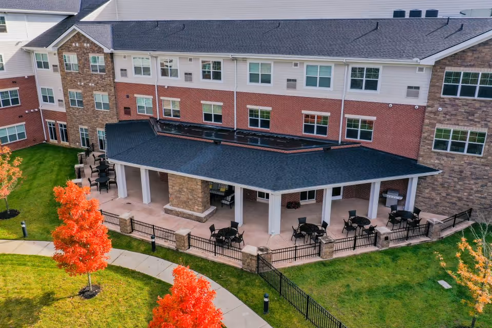 Outdoor patio area of a senior living facility with multiple round tables and chairs under a covered roof attached to a multi-story brick and stone building. The patio is surrounded by a black metal fence and landscaped with green grass and trees with bright red autumn leaves. A curved sidewalk runs in front of the patio.
