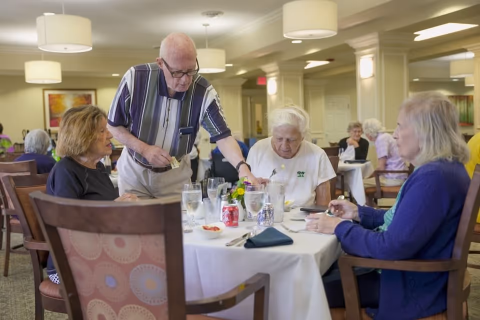 A group of elderly people sitting around a dining table in a senior living facility. One man is standing and appears to be serving or assisting the seated women. The table is set with glasses of water, soda cans, and utensils. The room is well-lit with ceiling lights and has other people dining in the background.