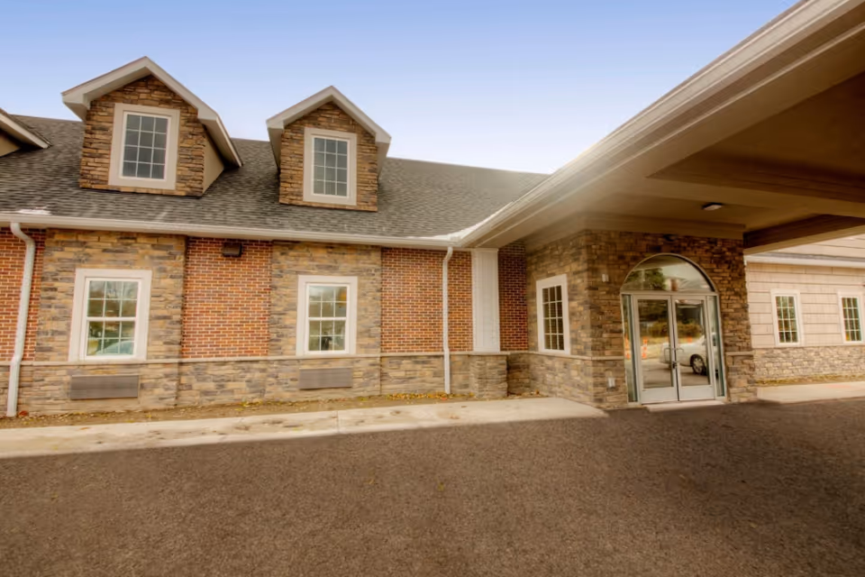 Front entrance of a brick-and-stone assisted living building with dormer windows and a covered glass-door entryway.