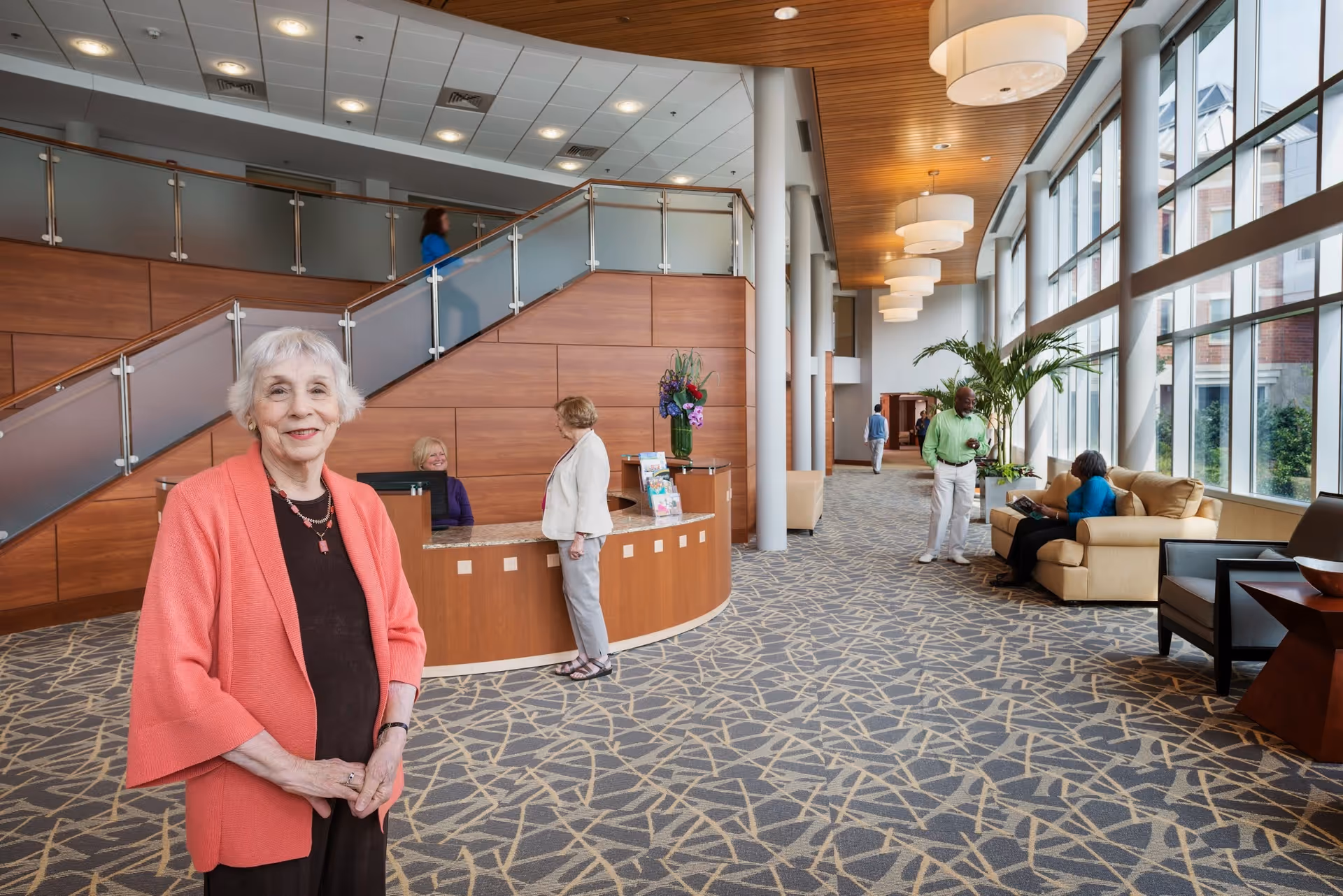 A spacious and well-lit lobby area of a retirement community with large windows on the right side letting in natural light. An elderly woman in a coral cardigan stands smiling in the foreground. Behind her is a curved wooden reception desk with two women interacting. A man and a woman are conversing near a beige sofa set along the windows. The area features a patterned carpet, modern ceiling lights, and a staircase with glass and metal railings on the left side.
