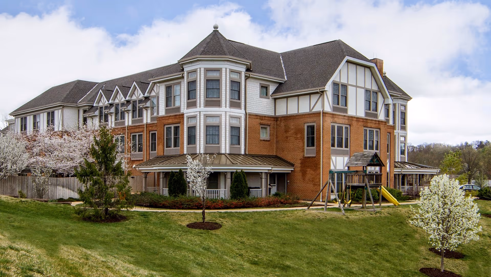 Exterior view of a multi-story senior living facility building with a combination of brick and white siding. The building has multiple windows and a pitched roof. In front of the building, there is a grassy area with small trees and a playground set with a slide and swings. The sky is partly cloudy.