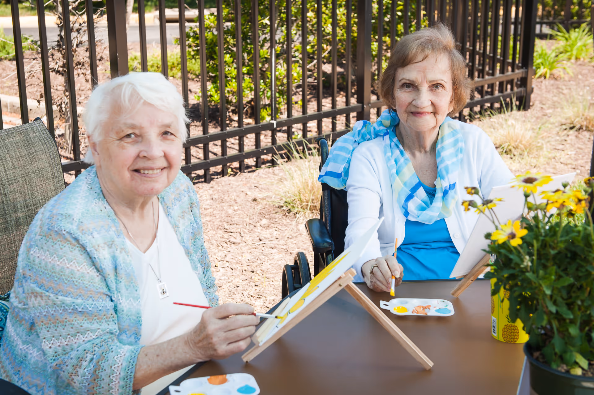 Two elderly women sitting outdoors at a table, painting on canvases with paint palettes in front of them. One woman has white hair and is wearing a light blue patterned cardigan, while the other woman has light brown hair and is wearing a blue top with a white cardigan and a blue scarf. There is a black metal fence and greenery in the background.