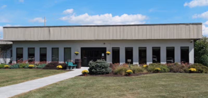 Front exterior of a single-story healthcare building with a central entrance, walkway, lawn, and landscaped shrubs.